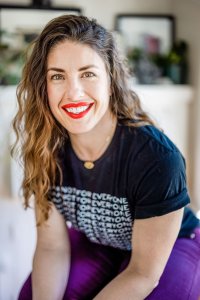Paleo to intuitive eating: brown haired women with black shirt, purple pants, red lipstick. She is sitting down and smiling.