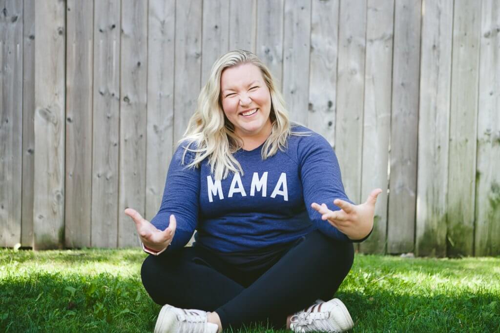Jenn Green, woman with blond hair, sitting cross legged with a blue "mama" shirt on and black pants. Jenn Green shares about post-partum mindset on the show!