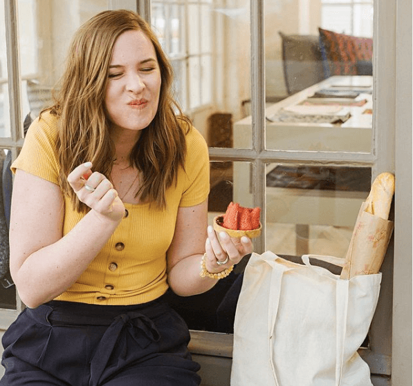 My dieting story {replay} Blond haired woman wearing a yellow shirt and black pants, sitting outdoors by a shop window and eating vibrant red strawberries. White canvas bag with French bread sits near her.